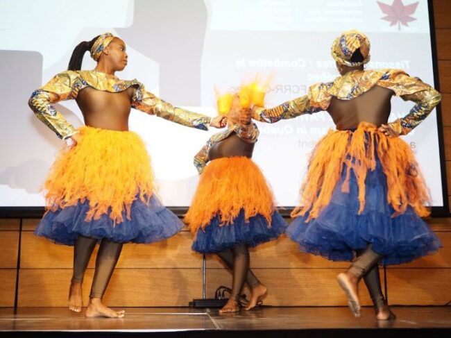 Three Black women dressed in yellow, dancing in a circle on stage at an event